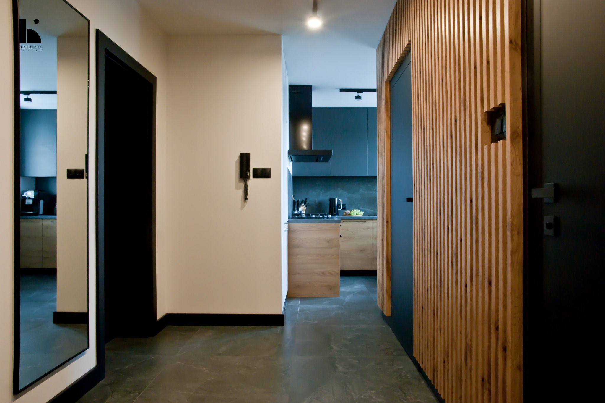 Nowoczesne wnętrze mieszkania w Łodzi: korytarz z lustrem, drewnianą ścianką działową i widokiem na kuchnię z czarnymi frontami i drewnianymi akcentami.