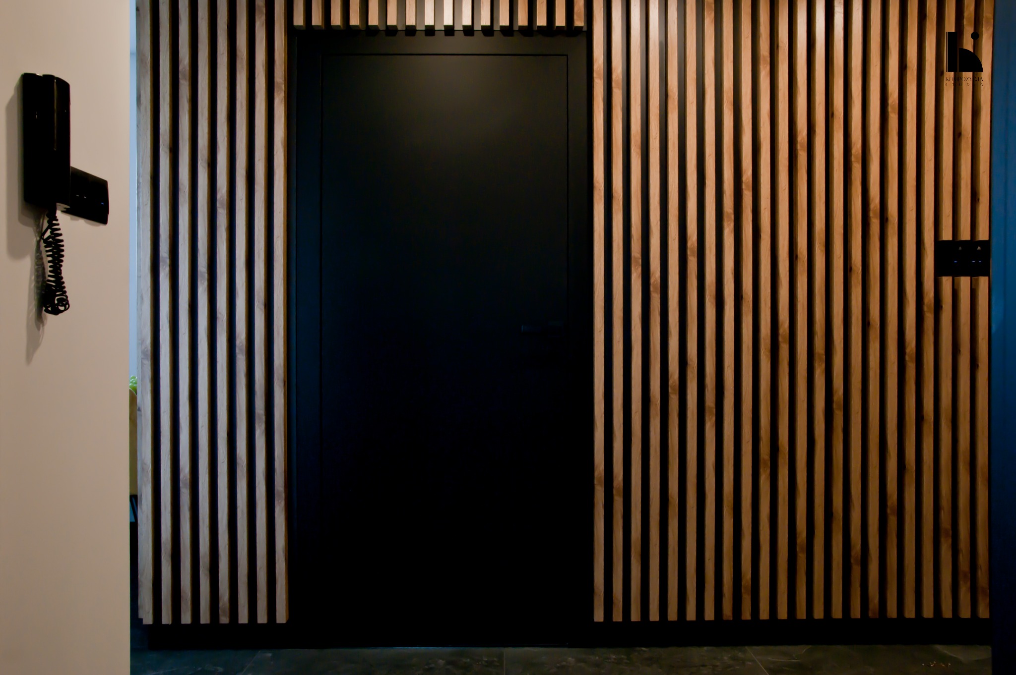 Nowoczesne wnętrze: czarne drzwi ukryte w drewnianej ściance z pionowych listew, czarny domofon na ścianie obok.