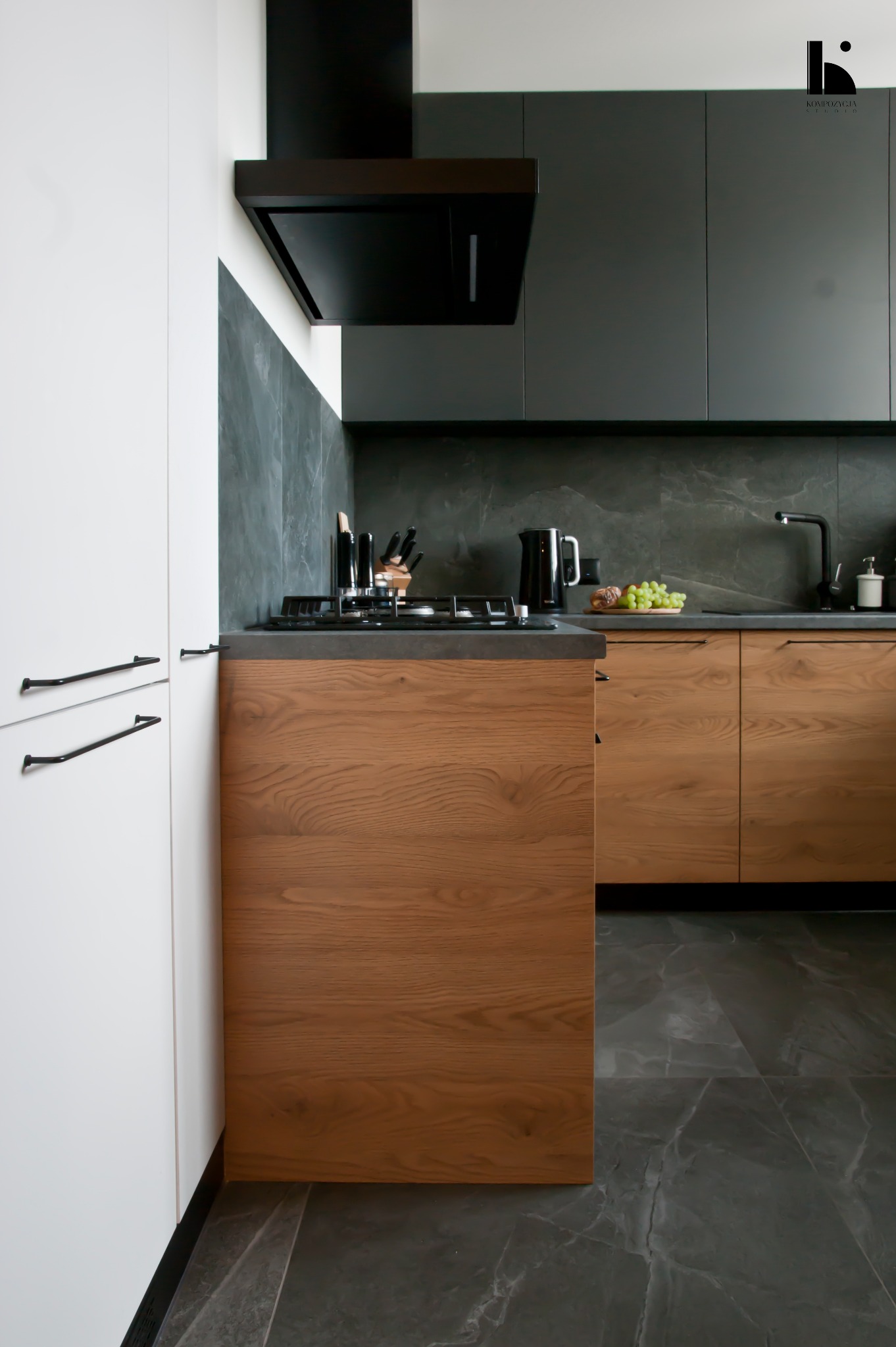 Nowoczesna kuchnia z drewnianymi frontami szafek, czarnymi elementami wykończenia i marmurową podłogą. Na blacie widoczne owoce i akcesoria kuchenne.