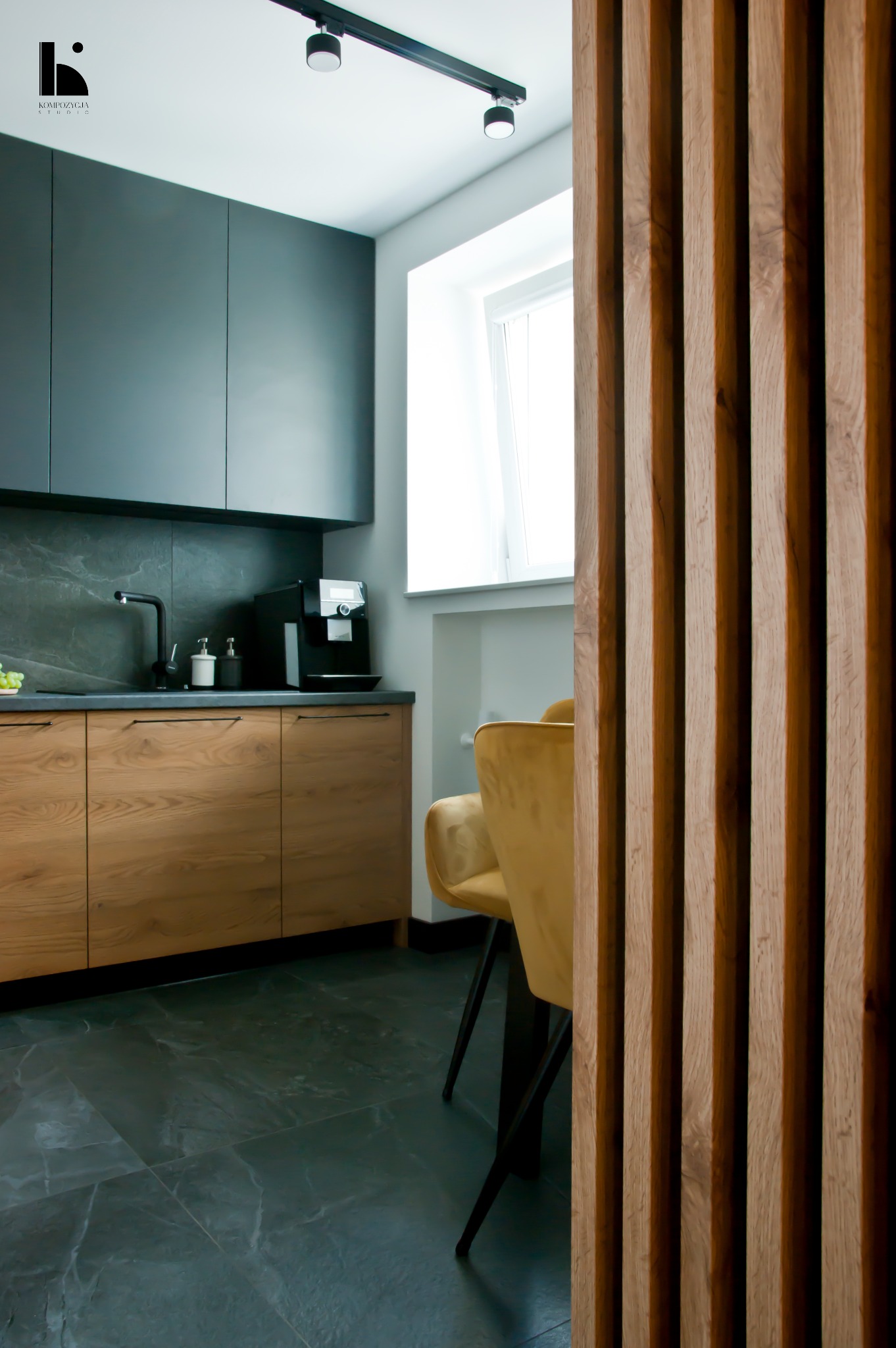 Nowoczesna kuchnia z drewnianymi szafkami, czarnym blatem i ścianą z płytek imitujących kamień, oddzielona od części jadalnej drewnianą ażurową ścianką. W tle widoczne krzesła w kolorze musztardowym.