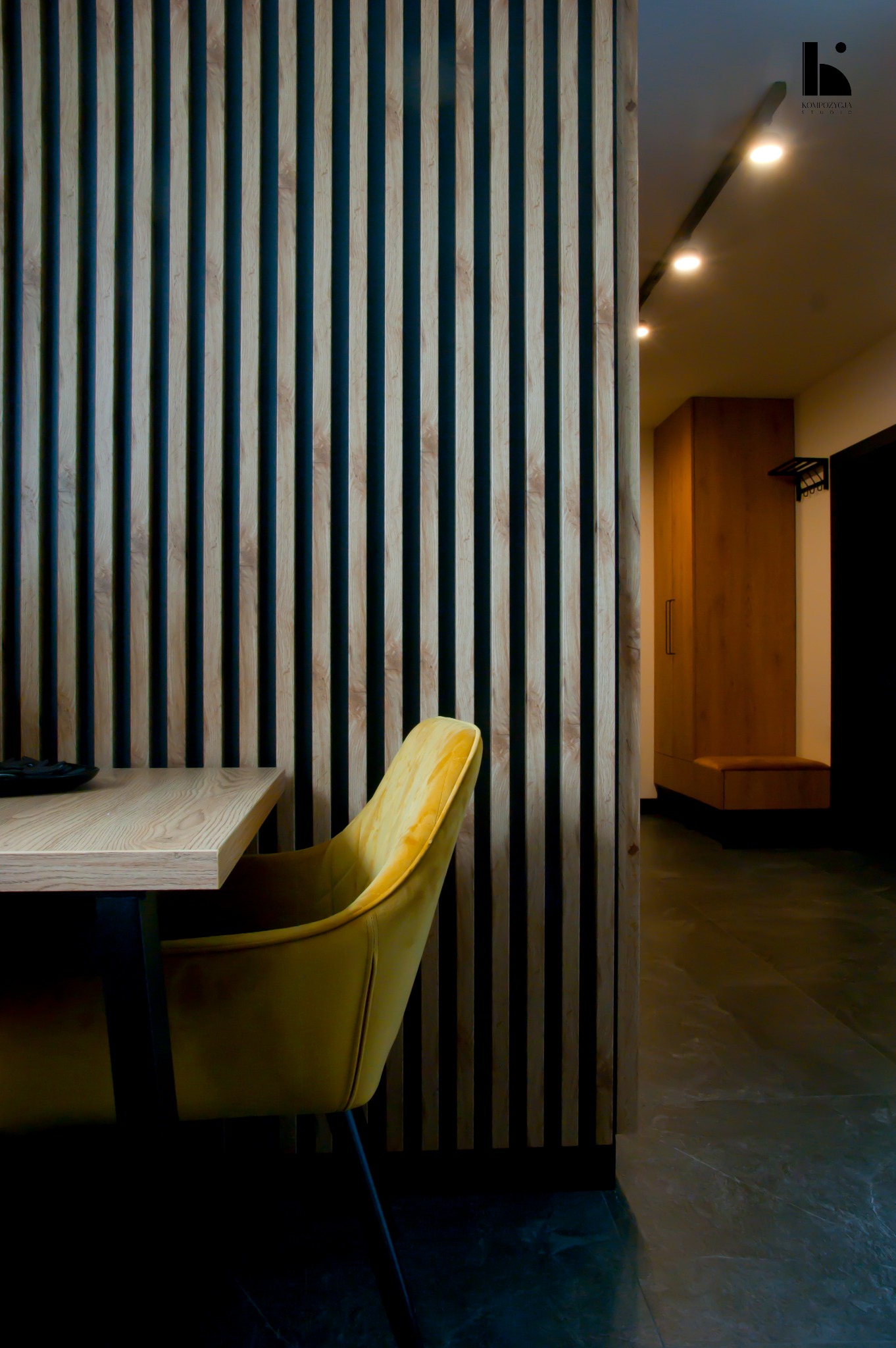 Nowoczesne wnętrze z drewnianymi lamelami oddzielającymi przestrzeń, żółte welurowe krzesło przy jasnym biurku, w tle widoczna szafa wnękowa i oświetlenie punktowe.