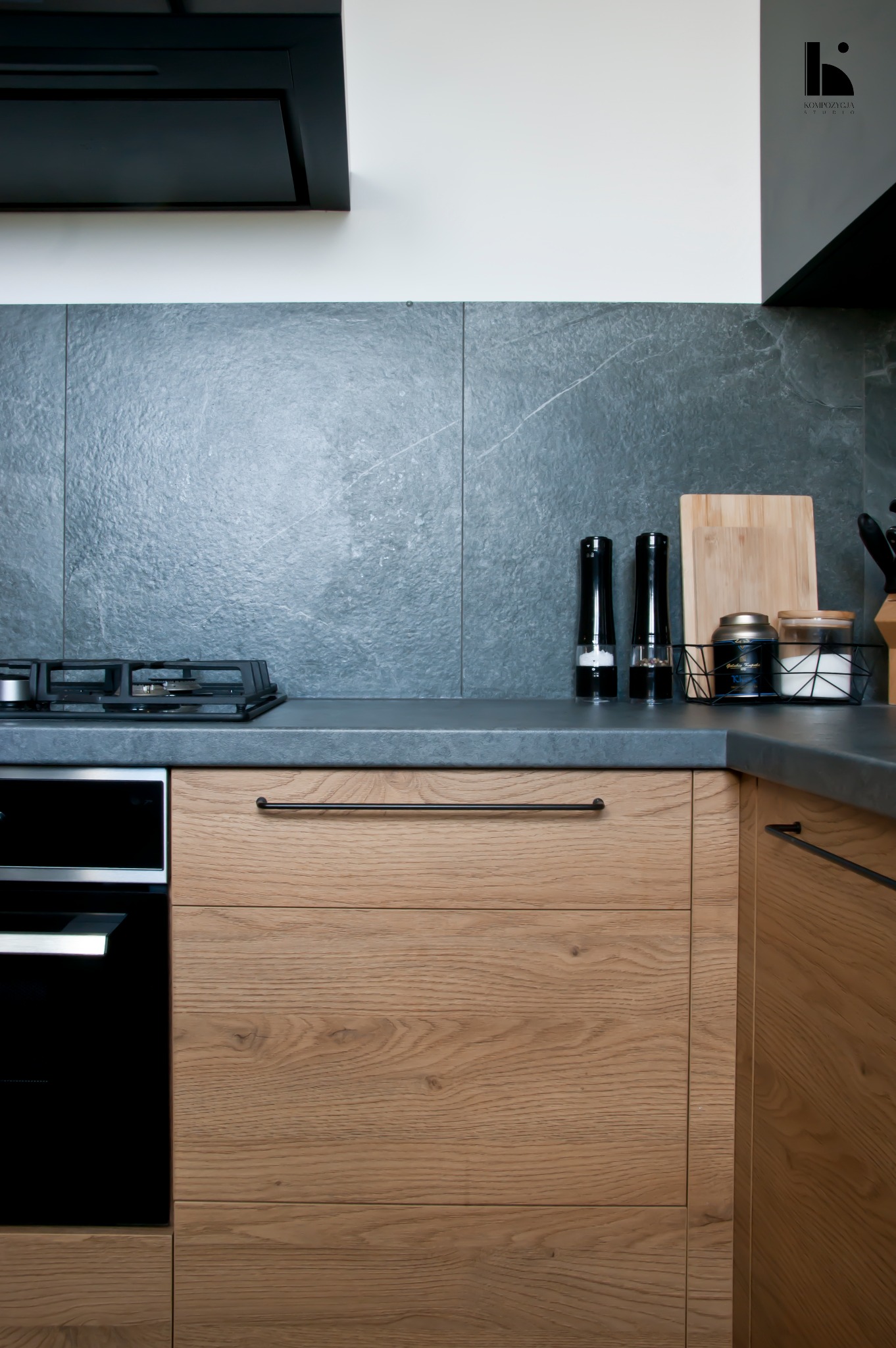 Fragment nowoczesnej kuchni z drewnianymi frontami szafek, szarym blatem i płytkami imitującymi kamień, widoczna płyta gazowa i akcesoria kuchenne, oświetlenie punktowe.