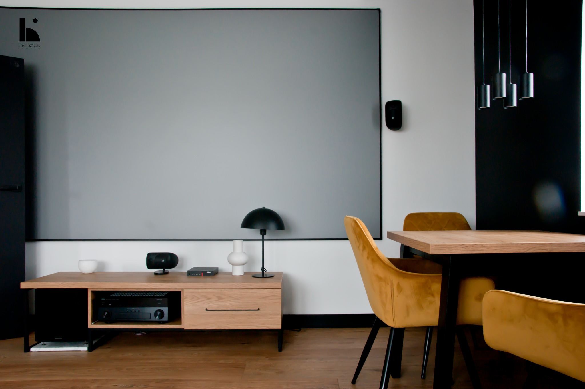 Jasne drewno i aksamitne krzesła w nowoczesnej aranżacji salonu z dużym ekranem projekcyjnym w Łodzi. Minimalistyczna komoda pod telewizorem z czarnymi akcentami i lampką.