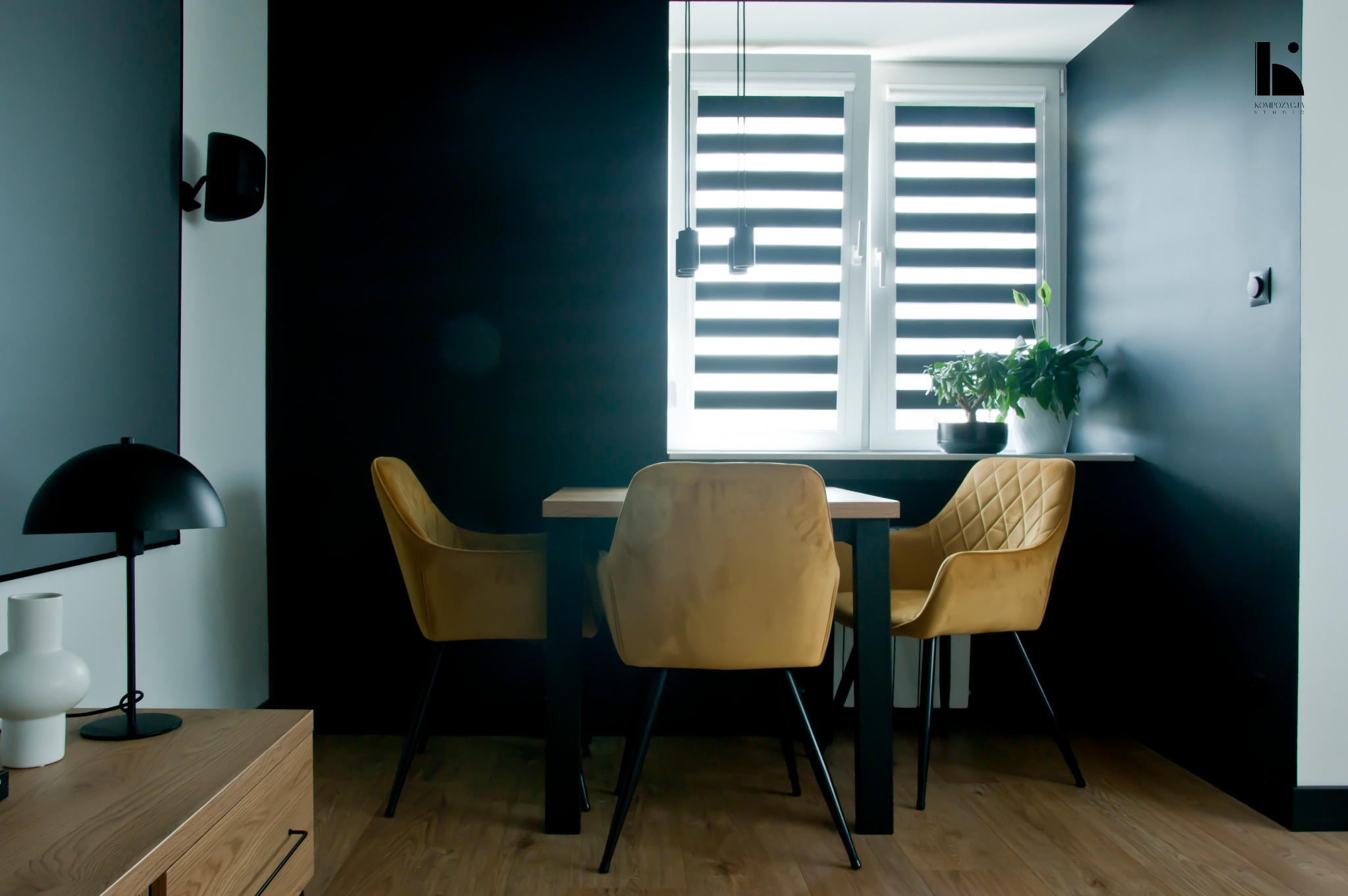 Nowoczesna jadalnia z ciemnymi ścianami, drewnianym stołem i musztardowymi krzesłami tapicerowanymi w romby, oświetlona przez okno z roletami dzień-noc i wiszące lampy.