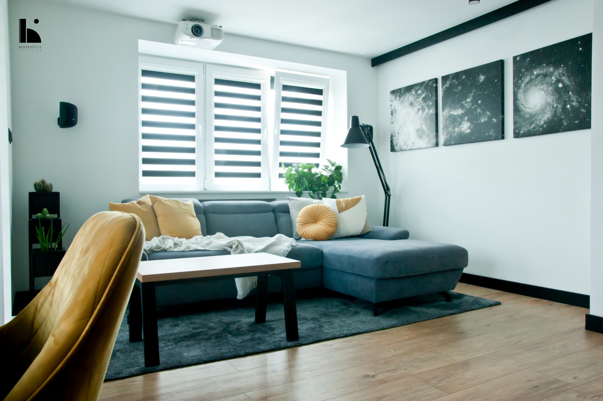 Nowoczesny salon z szarym narożnikiem, drewnianym stolikiem kawowym, trzema obrazami galaktyk na ścianie, projektorem na suficie i złotym fotelem na pierwszym planie.