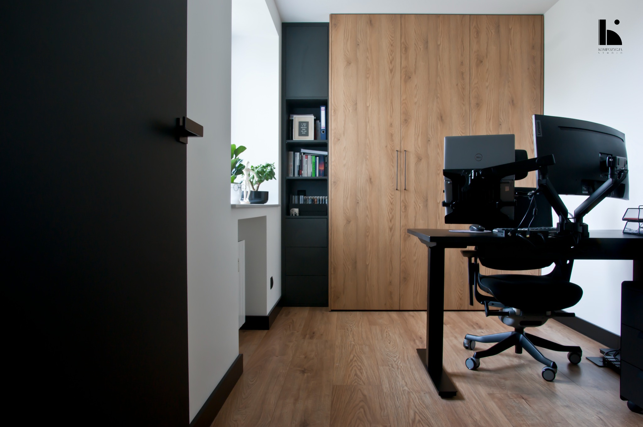 Nowoczesny gabinet z biurkiem, ergonomicznym krzesłem, dwoma monitorami na ramionach i szafą z drewnianymi frontami, uzupełniony o czarny regał z książkami i roślinami w oknie.