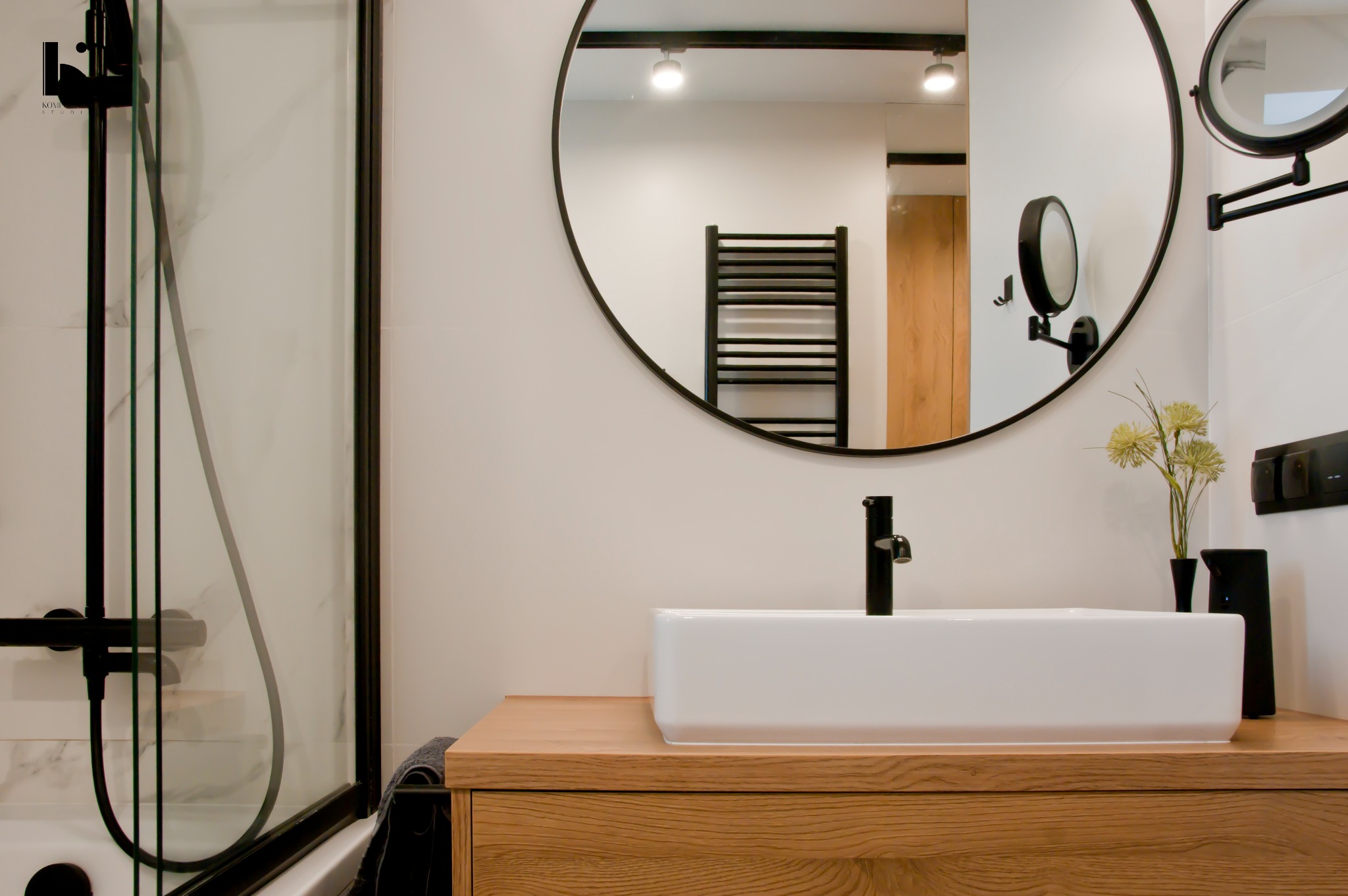 Nowoczesna łazienka z kabiną prysznicową, okrągłym lustrem z czarną ramą, białą prostokątną umywalką na drewnianej szafce i czarną armaturą; w odbiciu lustra widoczny grzejnik drabinkowy.