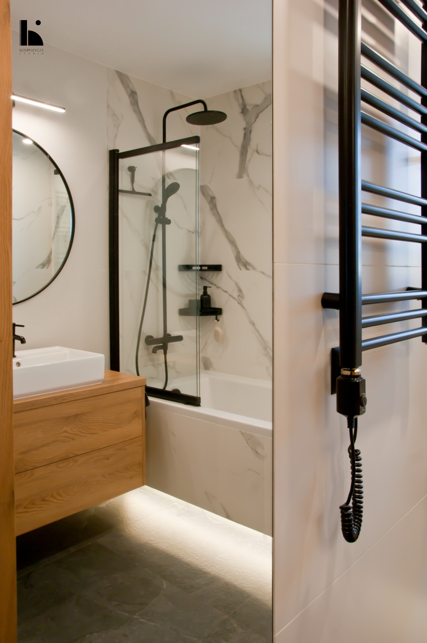 Nowoczesna łazienka z marmurowymi płytkami, czarną armaturą i podświetlaną szafką umywalkową z drewnianym frontem. Widoczny okrągły lustro i grzejnik drabinkowy w kolorze czarnym.