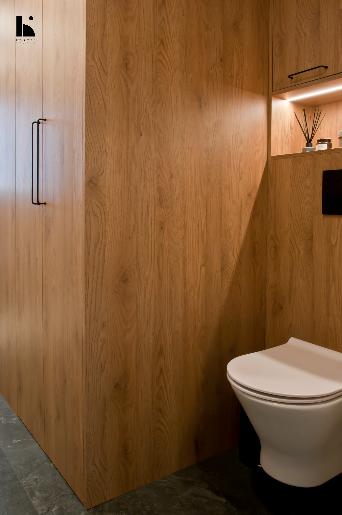 Nowoczesna łazienka z drewnianą zabudową, minimalistycznym WC i oświetloną półką z dekoracyjnymi elementami.