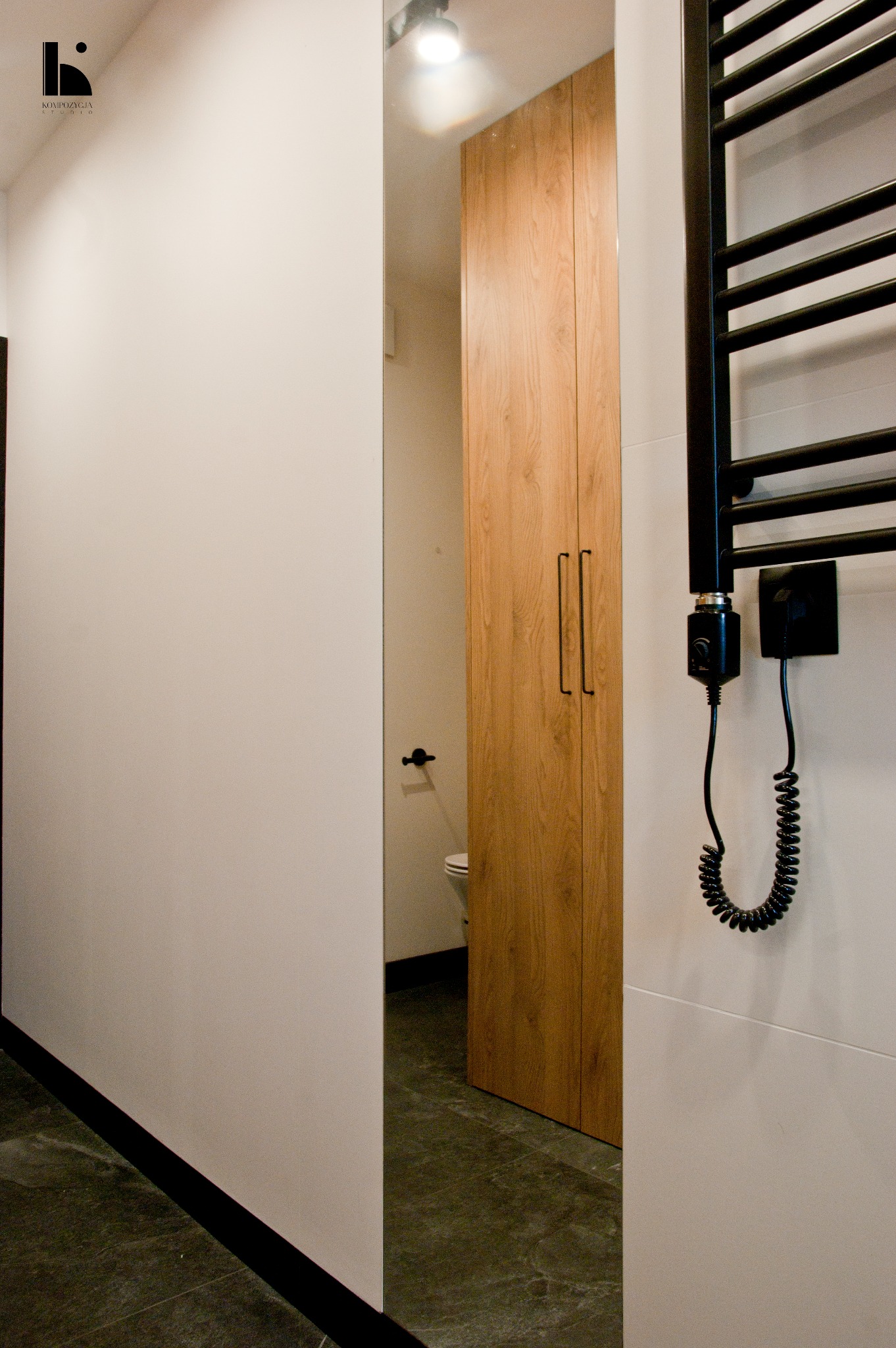 Nowoczesna łazienka z drewnianą szafką, czarnym grzejnikiem drabinkowym z elektrycznym zasilaniem i widoczną toaletą w głębi.