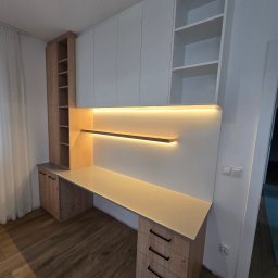 GAL - Nowoczesne biurko na wymiar z drewnianym blatem, oświetleniem LED pod szafkami wiszącymi i białymi frontami szafek górnych, z widoczną podłogą panelową i gniazdkami elektrycznymi na ścianie.