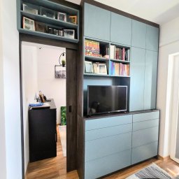 GAL - Niebiesko-brązowa zabudowa meblowa z szafkami, szufladami i półkami na książki, z wbudowanym telewizorem, widoczna z perspektywy podłogi, z otwartym przejściem do pokoju z biurkiem i lampą.