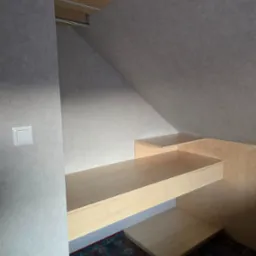 Półki i blat z jasnego drewna, zamontowane w skosie poddasza, z widocznym oświetleniem i wzorzystym dywanem na podłodze.