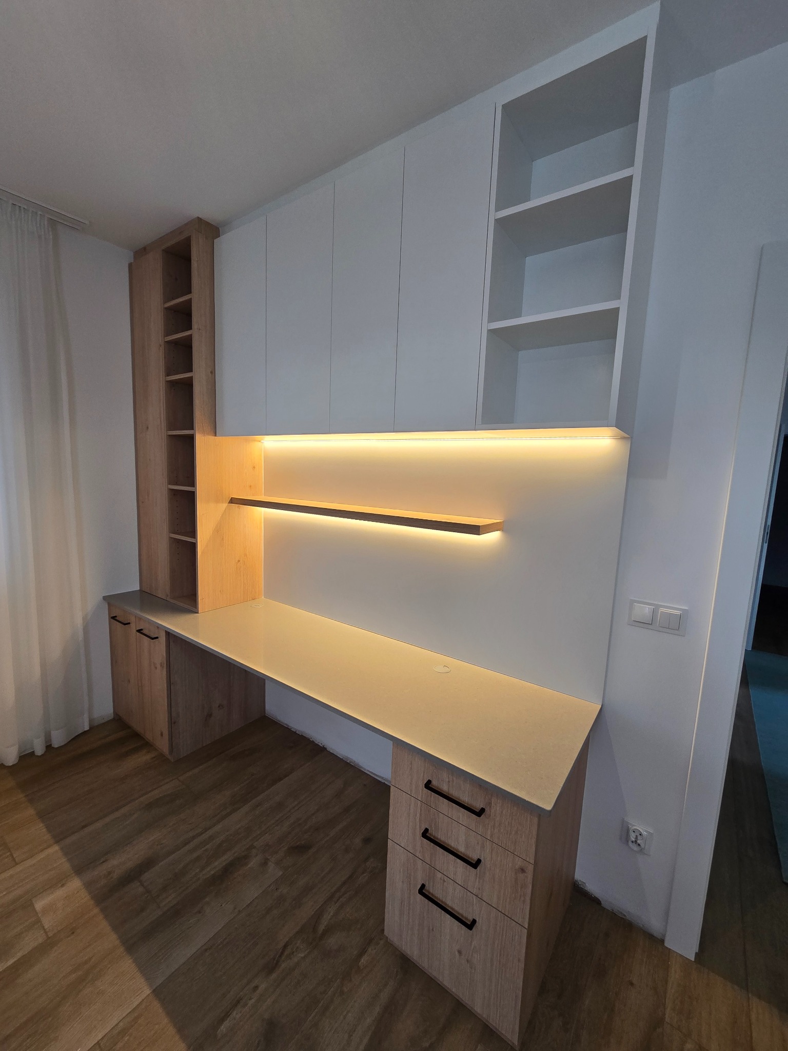 Nowoczesne biurko na wymiar z drewnianym blatem, oświetleniem LED pod szafkami wiszącymi i białymi frontami szafek górnych, z widoczną podłogą panelową i gniazdkami elektrycznymi na ścianie.