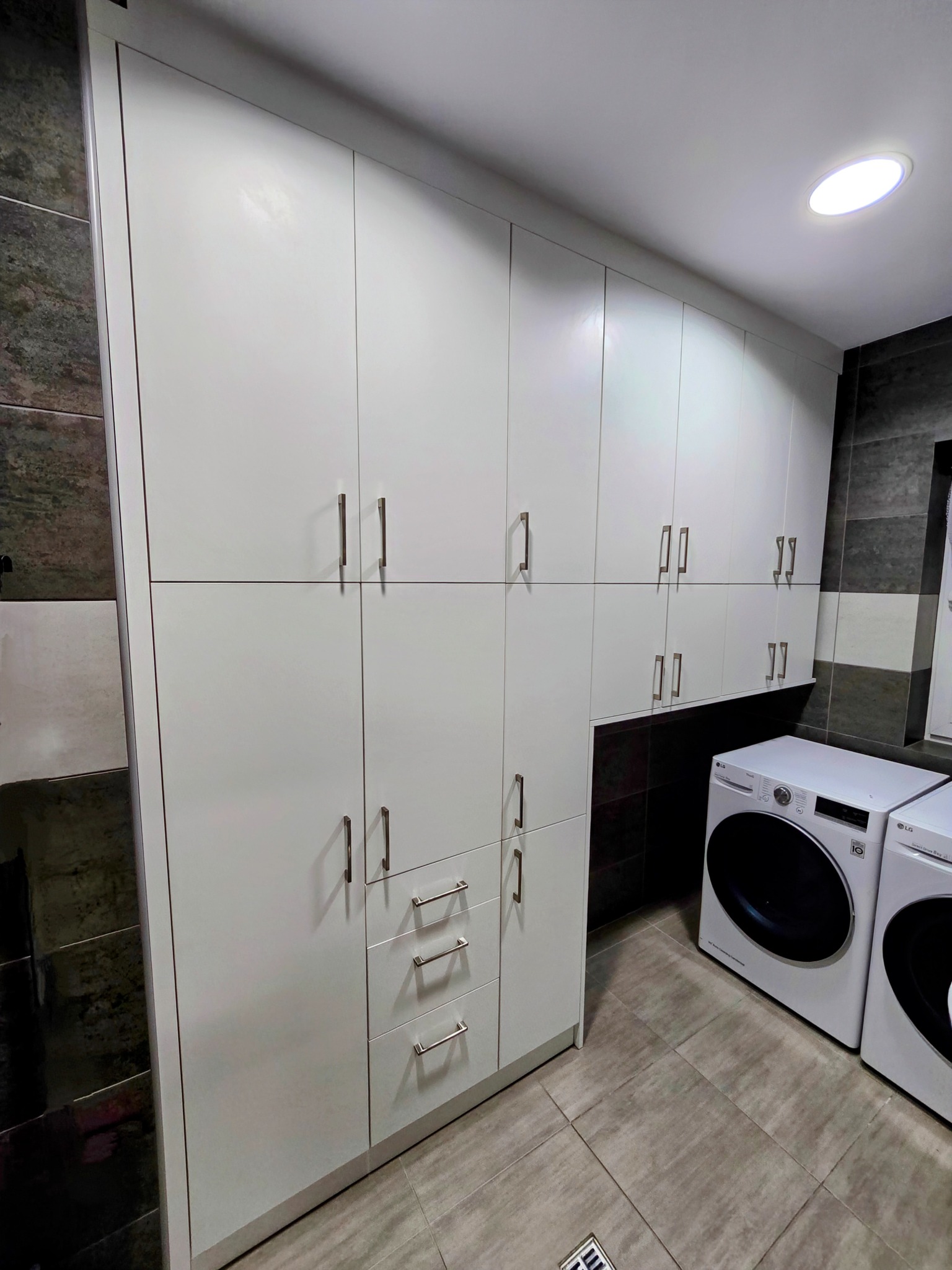 Białe szafy z szufladami i długimi uchwytami w pomieszczeniu gospodarczym z pralkami LG, szare płytki na podłodze i ścianach.