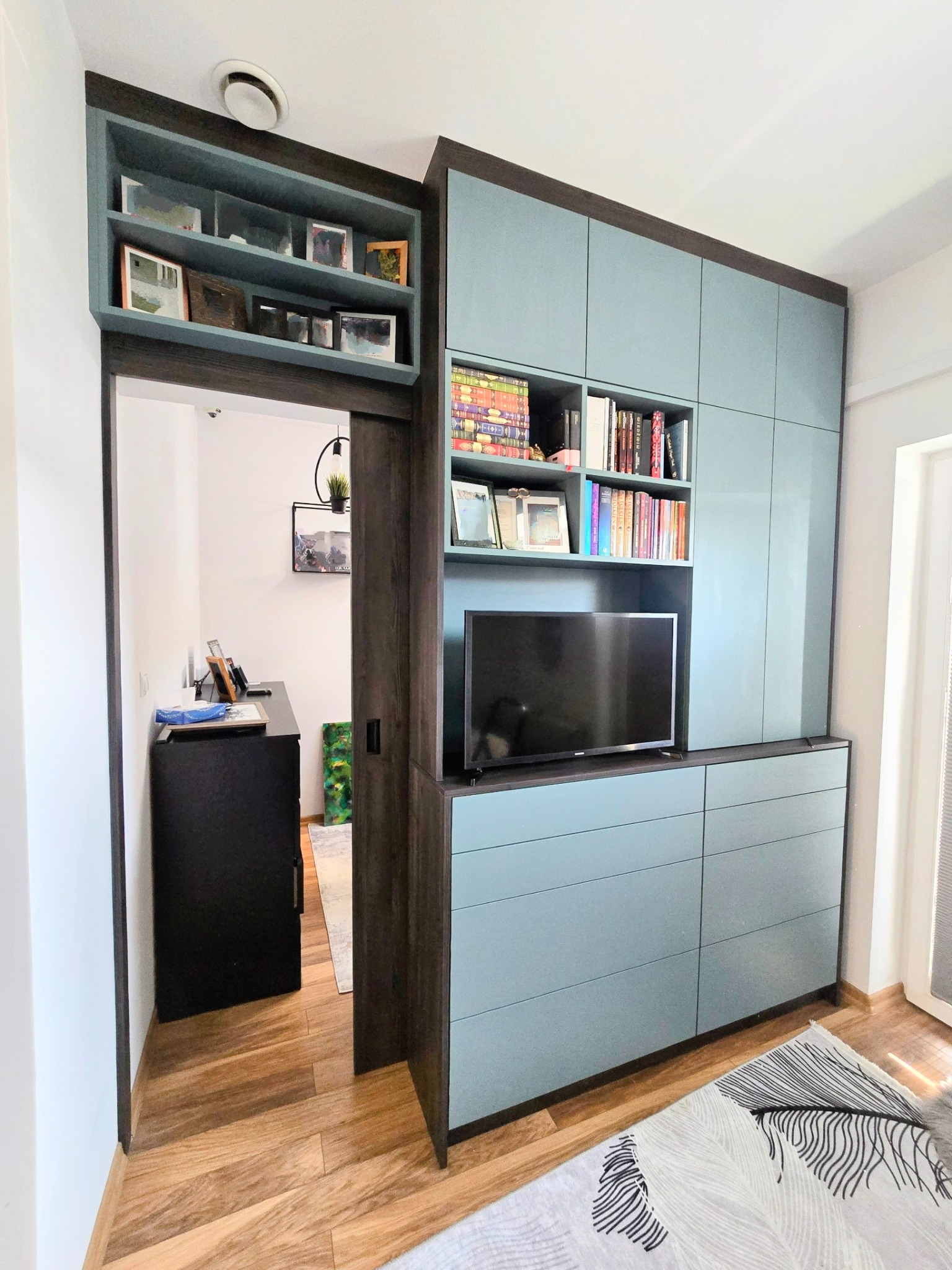 Niebiesko-brązowa zabudowa meblowa z szafkami, szufladami i półkami na książki, z wbudowanym telewizorem, widoczna z perspektywy podłogi, z otwartym przejściem do pokoju z biurkiem i lampą.