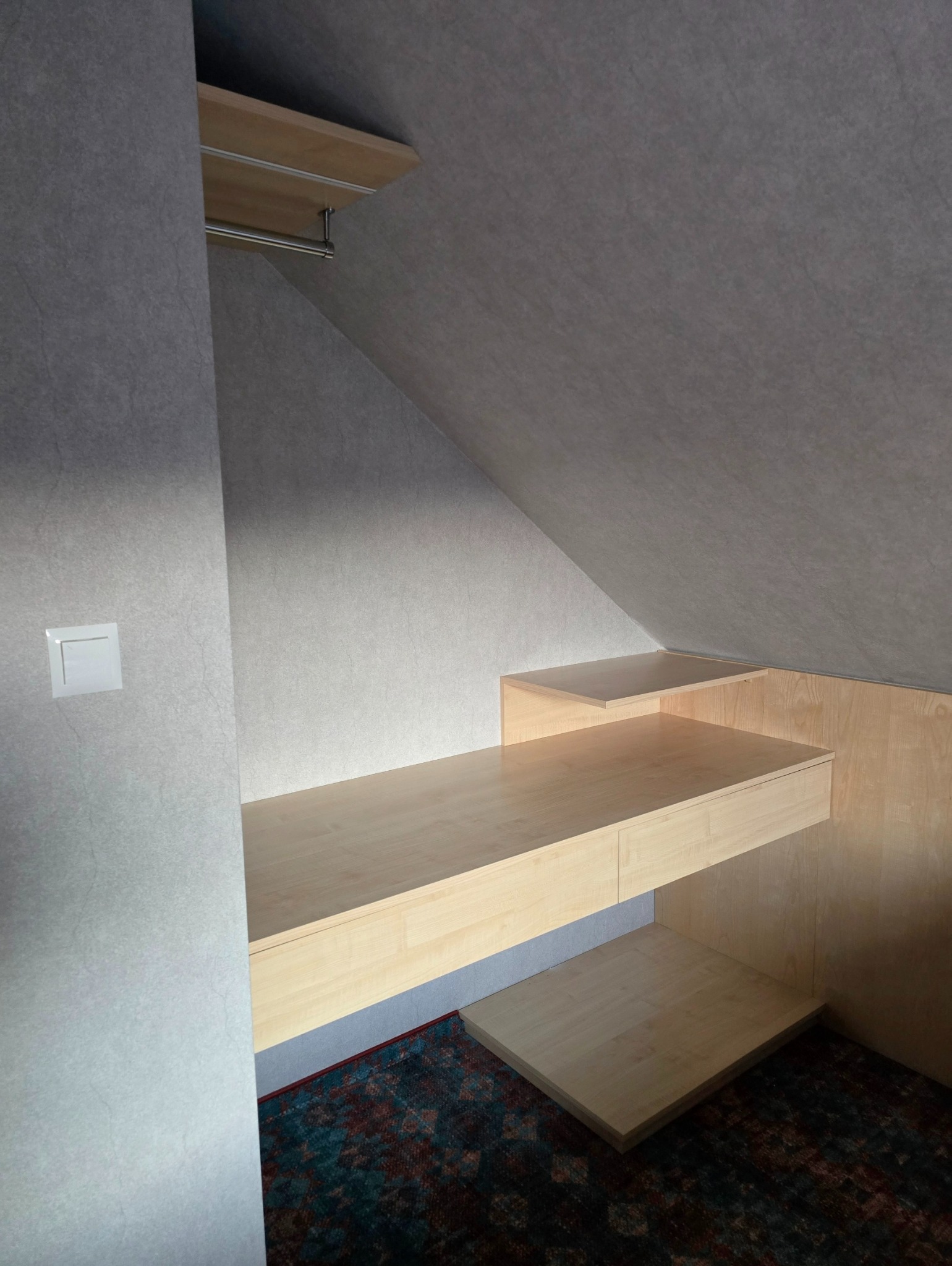 Półki i blat z jasnego drewna, zamontowane w skosie poddasza, z widocznym oświetleniem i wzorzystym dywanem na podłodze.