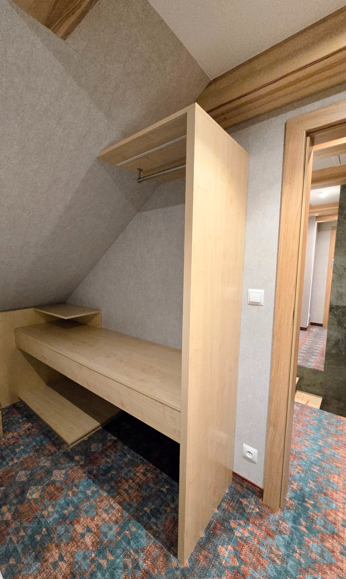 Jasna, drewniana szafa wnękowa z półkami i drążkiem na ubrania, wkomponowana w skos dachu, z widocznym dywanem w geometryczne wzory i fragmentem drzwi.