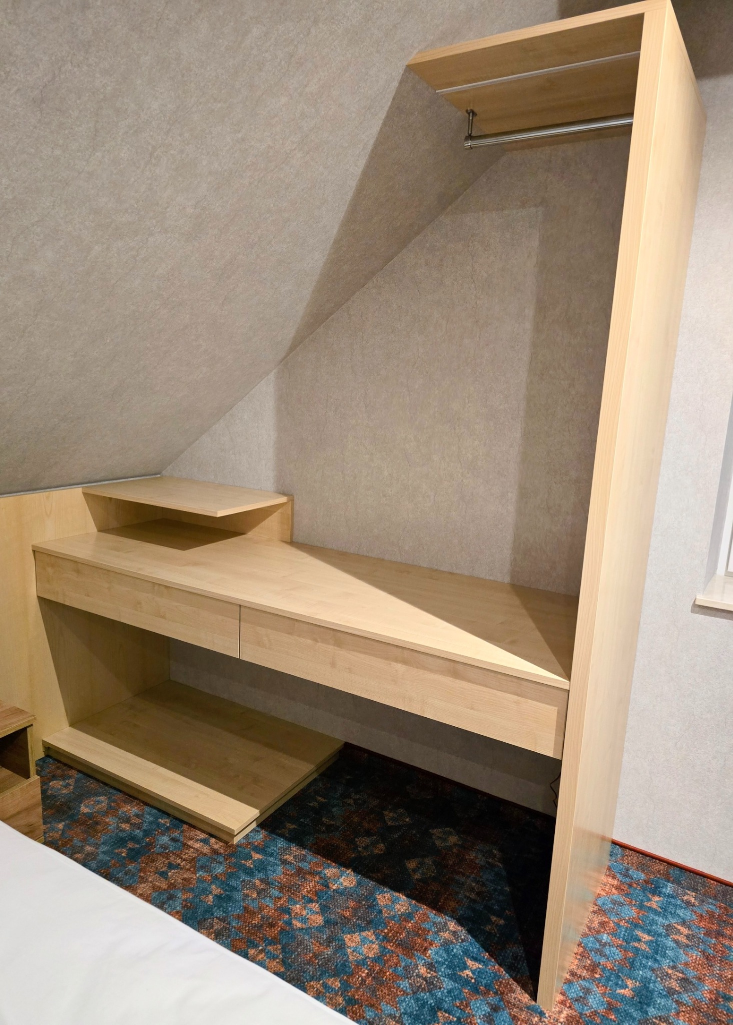 Jasna, drewniana szafa wnękowa z biurkiem i półkami, dopasowana do skosu dachu, z drążkiem na ubrania w górnej części, na tle wzorzystej wykładziny podłogowej.