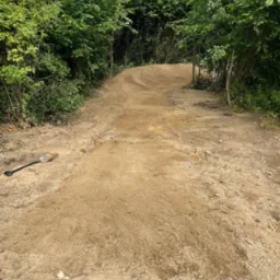 Ukształtowany teren z widoczną ścieżką z ziemi, otoczony zielenią drzew i krzewów, z łopatą leżącą na ziemi po lewej stronie.