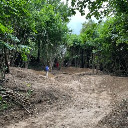 GRAVITY SKATE SHOP STANISŁAW UKLEJA - Przygotowanie terenu w lesie, widoczni dwaj pracownicy, jeden w niebieskiej koszuli, drugi w oddali, usypywanie ziemi, wycinka drzew i krzewów, ślady opon na ziemi.