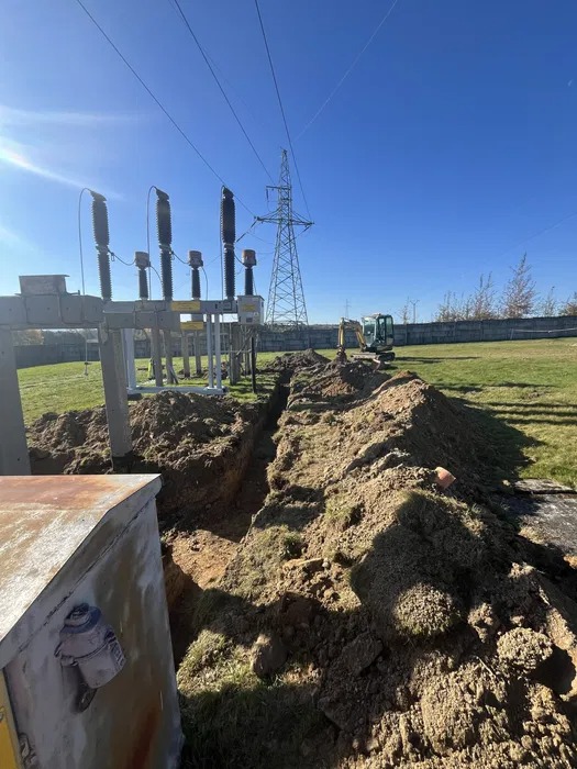 Wąski wykop w ziemi prowadzony wzdłuż infrastruktury elektrycznej, w tle widoczna wieża wysokiego napięcia i mała koparka.