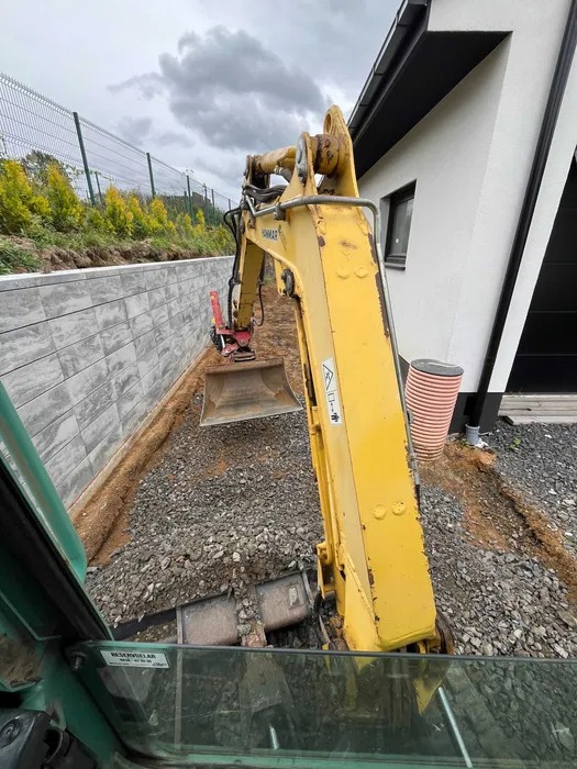 Widok z kabiny na żółtą mini-koparkę Yanmar podczas prac ziemnych, widoczny wykop z kamieniami, mur oporowy z betonowych płyt i fragment nowoczesnego domu.