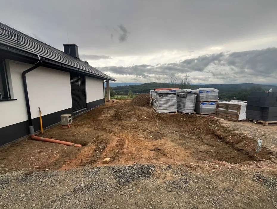 Przygotowanie terenu przy nowoczesnym domu: wykop pod instalacje, rura kanalizacyjna, materiały budowlane na paletach, widok na pagórkowaty krajobraz w pochmurny dzień.