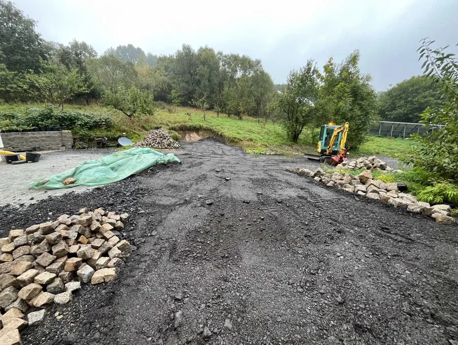 Przygotowanie podłoża pod drogę: wyrównany teren z wysypanym kruszywem, obrzeża z kamiennej kostki, mini koparka w tle.