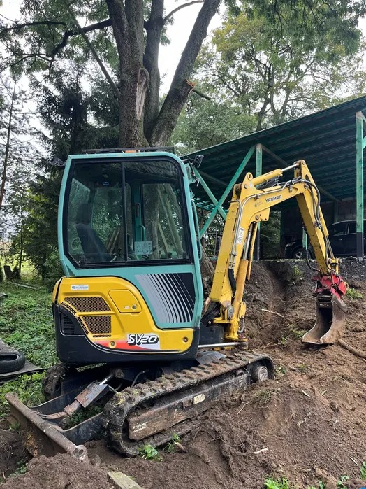 Żółto-turkusowa mini koparka Yanmar SV20 na gąsienicach, z czerwonym osprzętem do wykopów, stoi na świeżo usypanej ziemi pod dużym drzewem z widocznym ubytkiem kory.