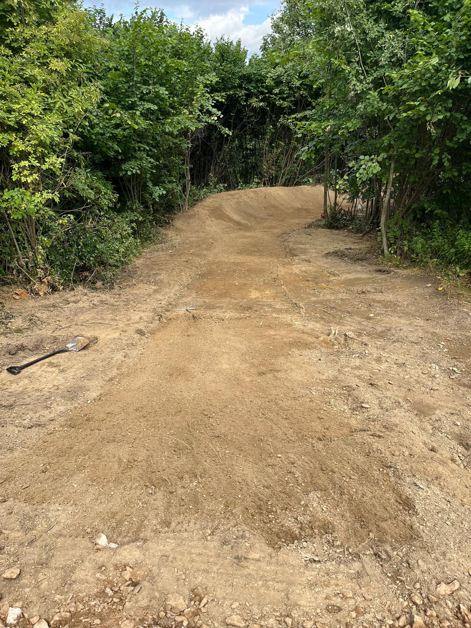 Ukształtowany teren z widoczną ścieżką z ziemi, otoczony zielenią drzew i krzewów, z łopatą leżącą na ziemi po lewej stronie.