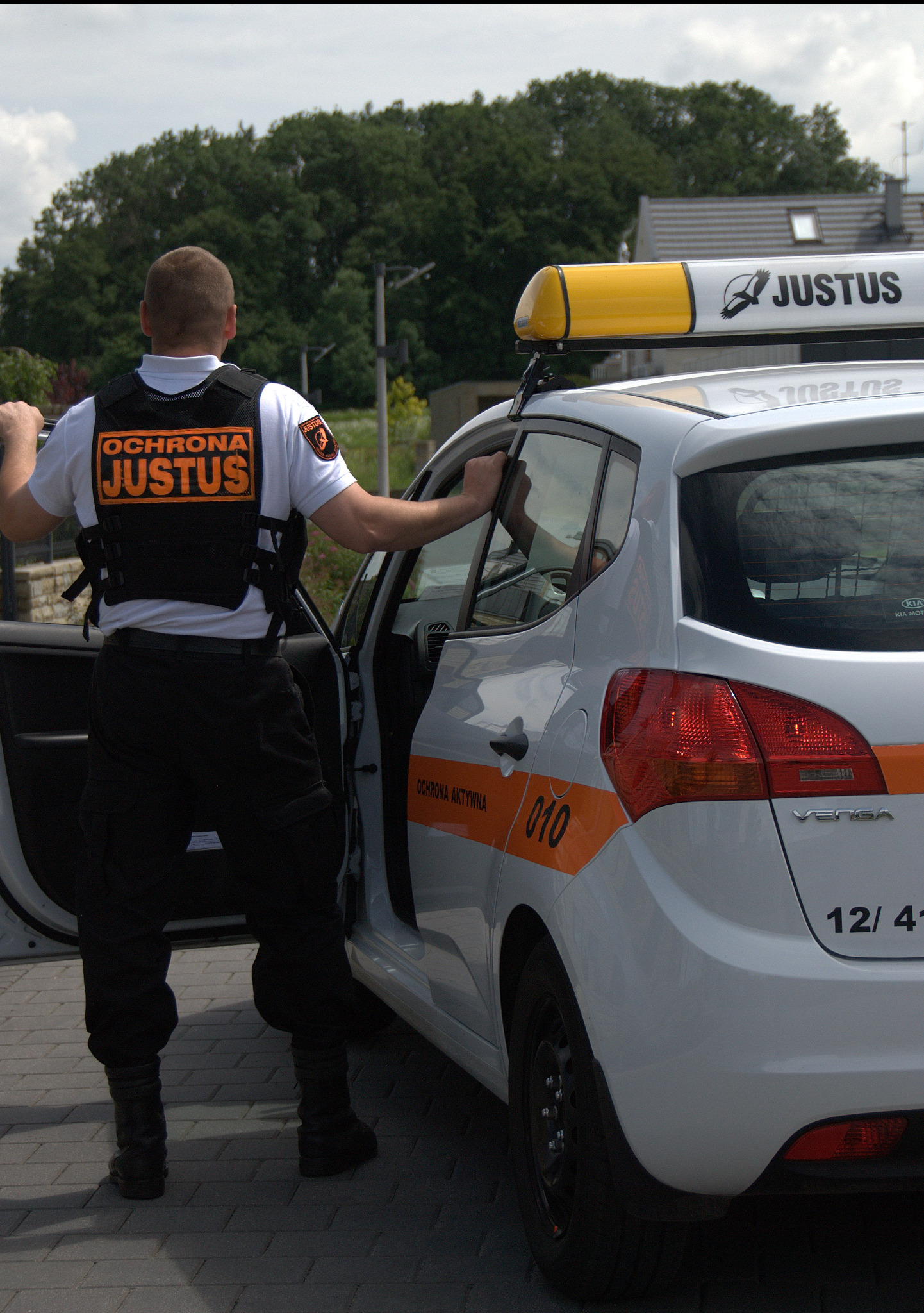 Ochroniarz w kamizelce firmy Justus otwiera drzwi białego samochodu z napisem 'Ochrona Aktywna 010' i logo Justus na dachu, widok z tyłu.