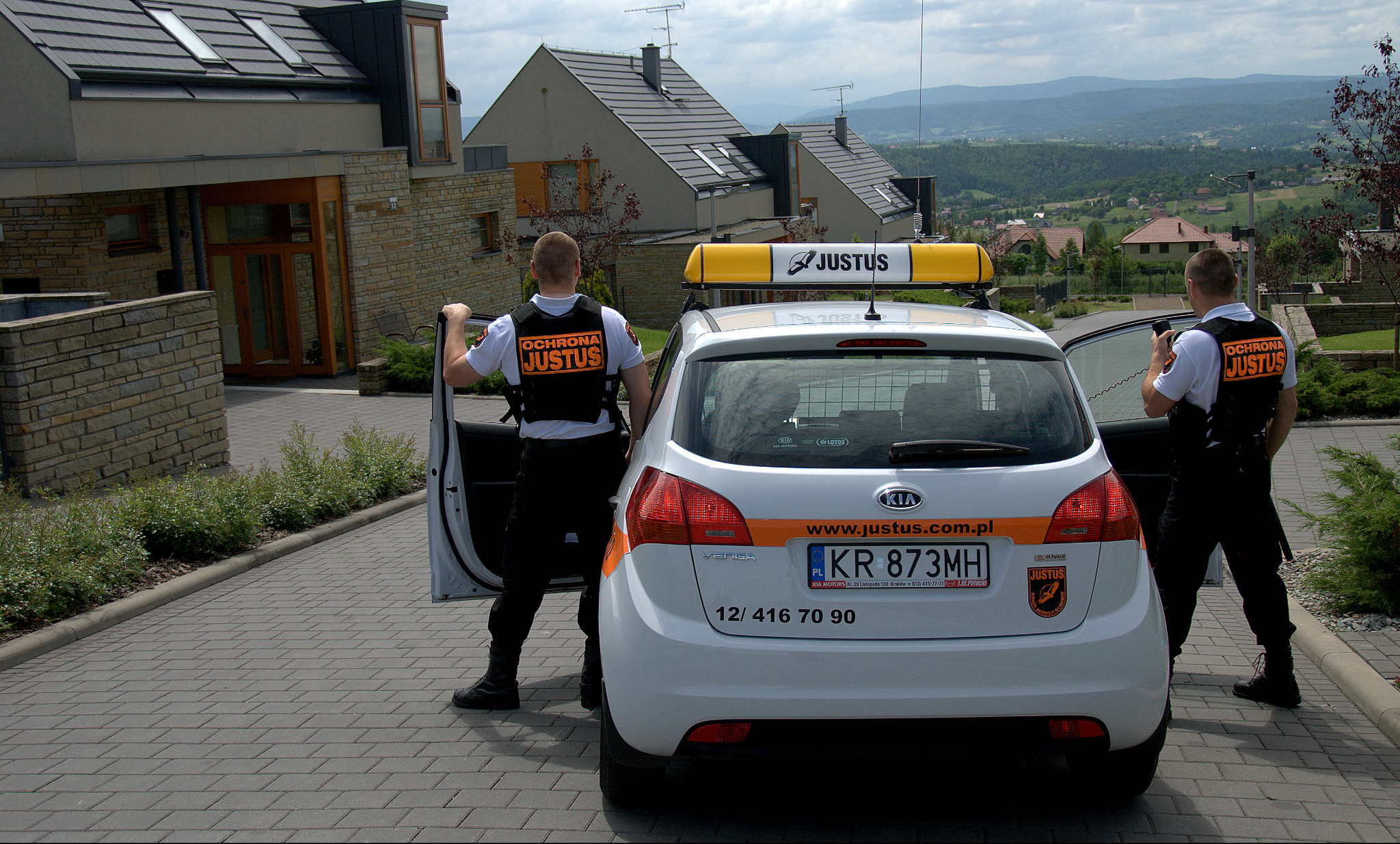 Samochód firmy ochroniarskiej Justus na tle nowoczesnego domu i gór. Dwóch ochroniarzy w uniformach stoi obok pojazdu. Widoczne logo firmy na samochodzie i kamizelkach.