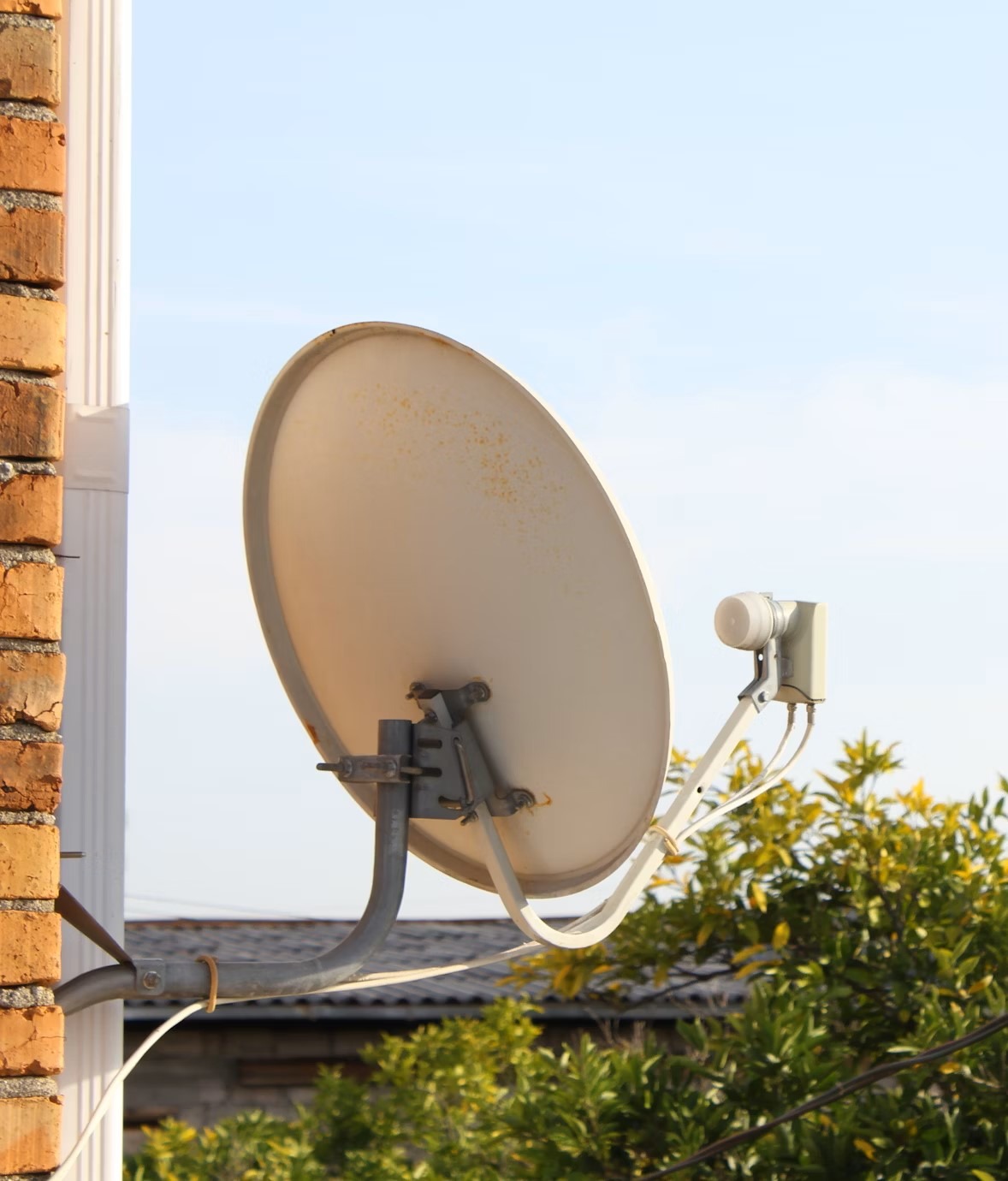 Zamontowana antena satelitarna przymocowana do ceglanej ściany budynku, na tle błękitnego nieba i zieleni. Widoczne kable i elementy mocujące.