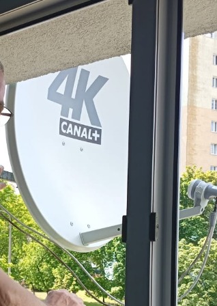 Biała antena satelitarna 4K Canal+ zamontowana na balkonie, widok zza okna na zieleń i budynek. Instalator widoczny po lewej stronie kadru.