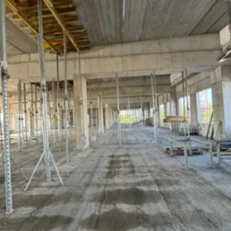 Wnętrze surowego, betonowego budynku w trakcie budowy, z widocznymi podporami stropowymi i deskowaniem, surowy beton na podłodze z widocznymi śladami.