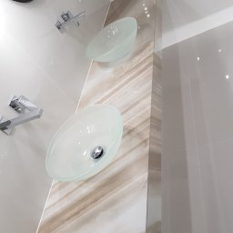 SMOLENSKI - Dwie szklane umywalki nablatowe z chromowanymi bateriami na ścianie wyłożonej beżowymi płytkami i dekoracyjnym panelem imitującym marmur. Nowoczesna łazienka.