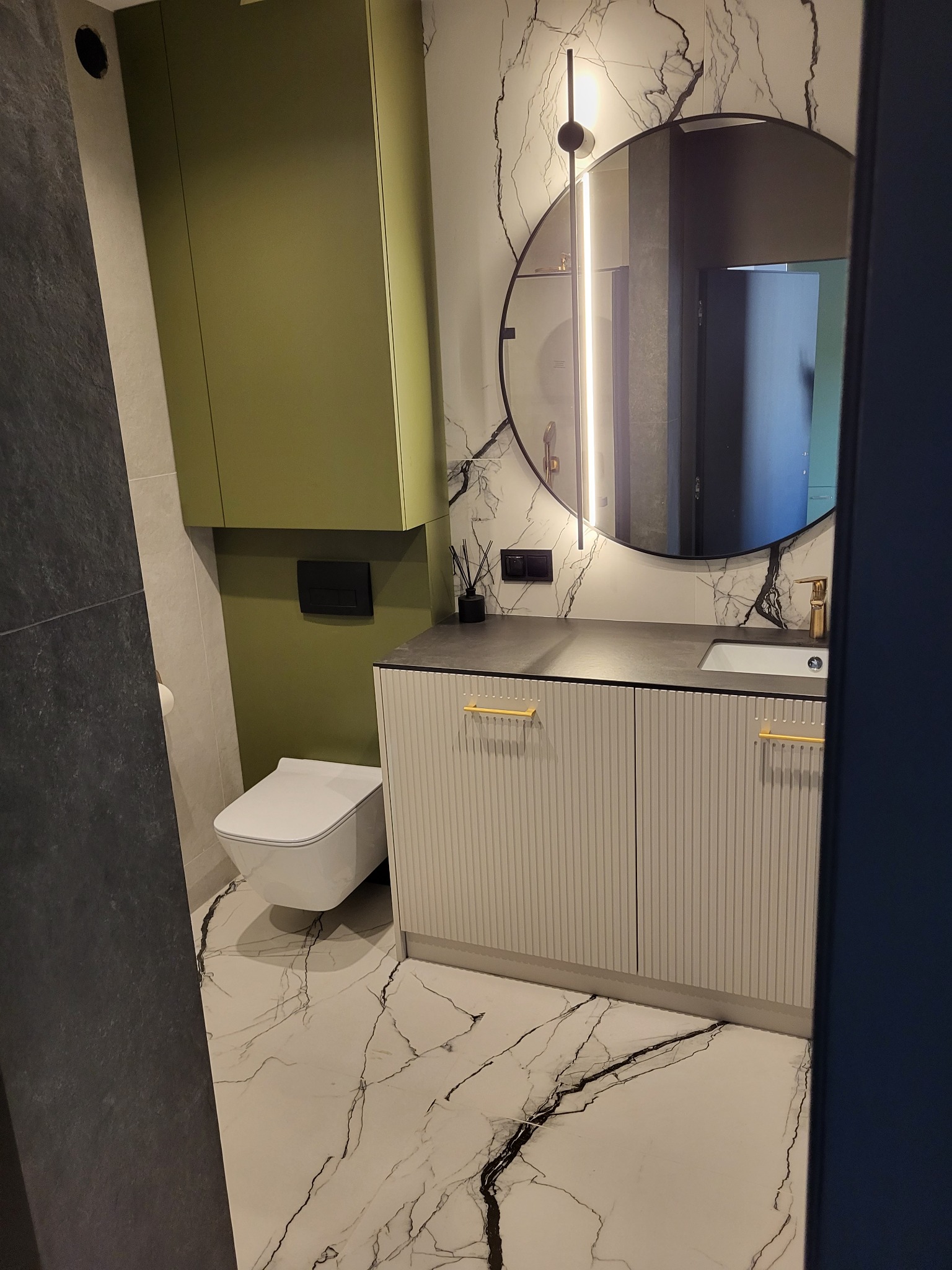 Nowoczesna łazienka z szafką pod umywalką w prążki, okrągłym lustrem z podświetleniem i wiszącą toaletą. Marmurowe płytki podłogowe z wyraźnym wzorem.