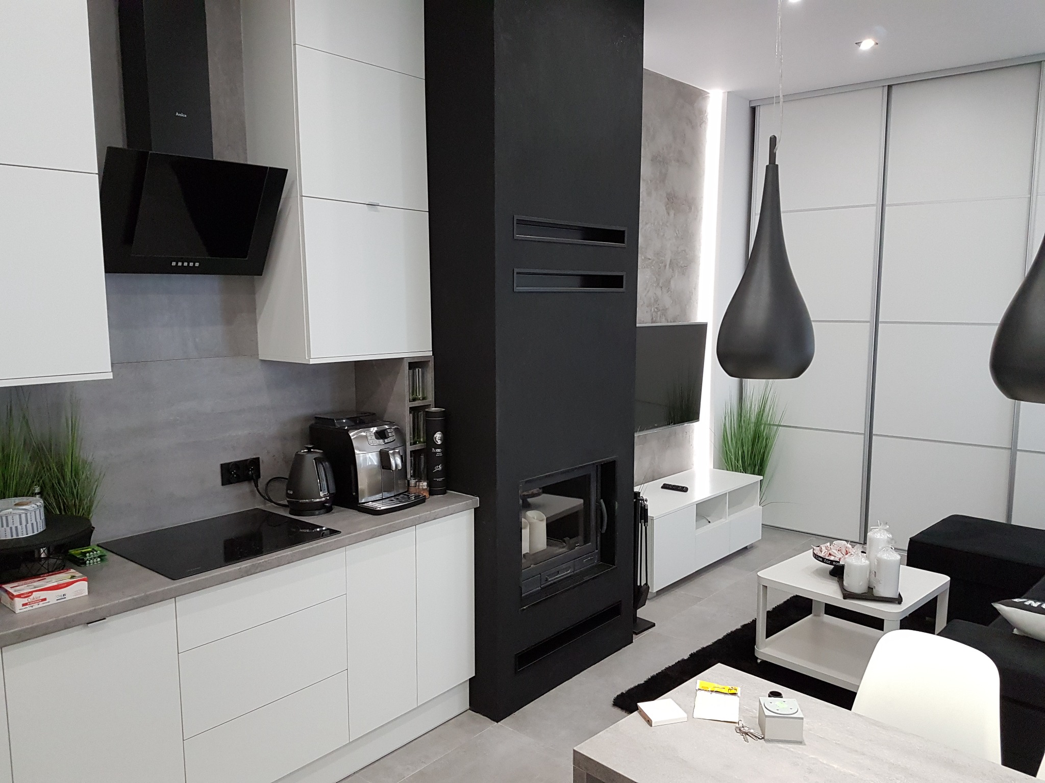 Nowoczesna kuchnia z czarnym kominkiem i białymi szafkami. Minimalistyczny design, szare ściany i podłogi. Oświetlenie punktowe i wiszące lampy.