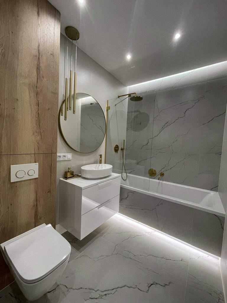 Nowoczesna łazienka z marmurową podłogą i ścianami, białą armaturą, złotymi akcentami i oświetleniem LED pod wanną. Okrągłe lustro i wisząca lampa dodają elegancji.