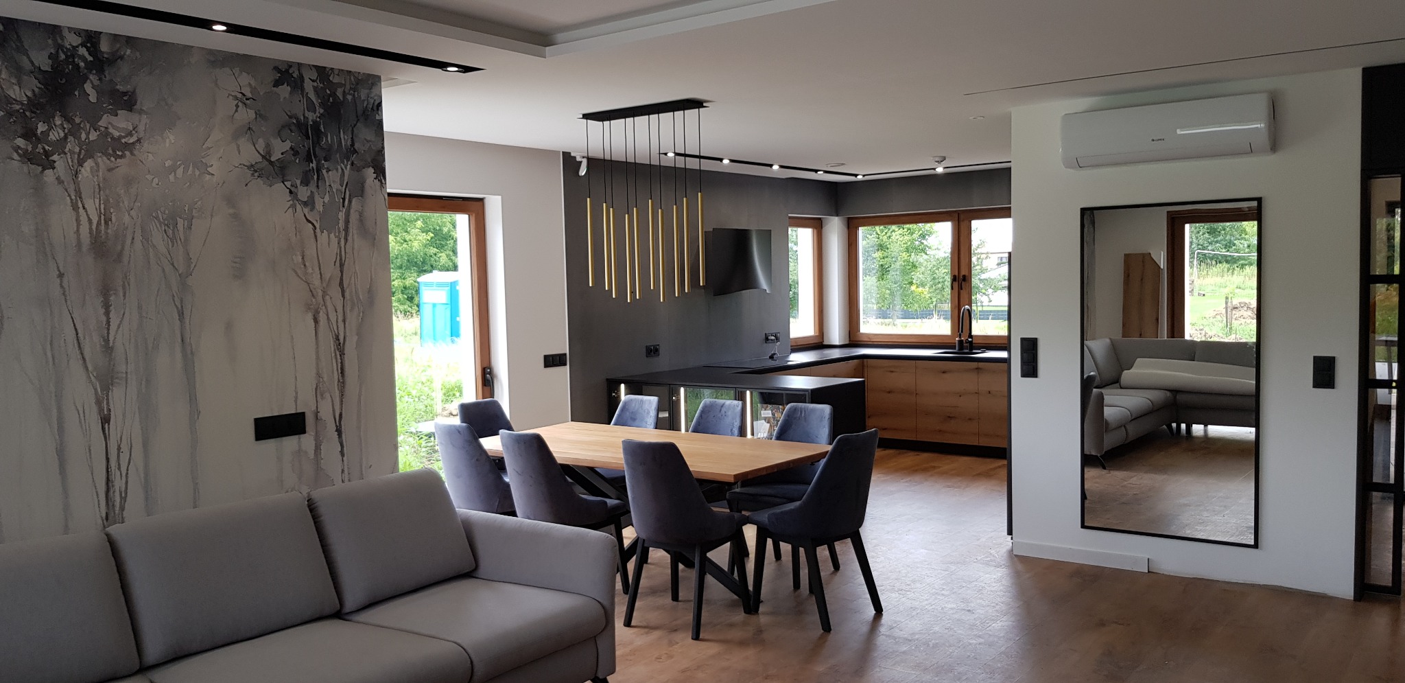 Nowoczesne wnętrze domu: szara sofa, stół z krzesłami, kuchnia z drewnianymi szafkami, czarny okap i lampa. Na ścianie dekoracyjny wzór drzew. Widok na zieleń za oknem.