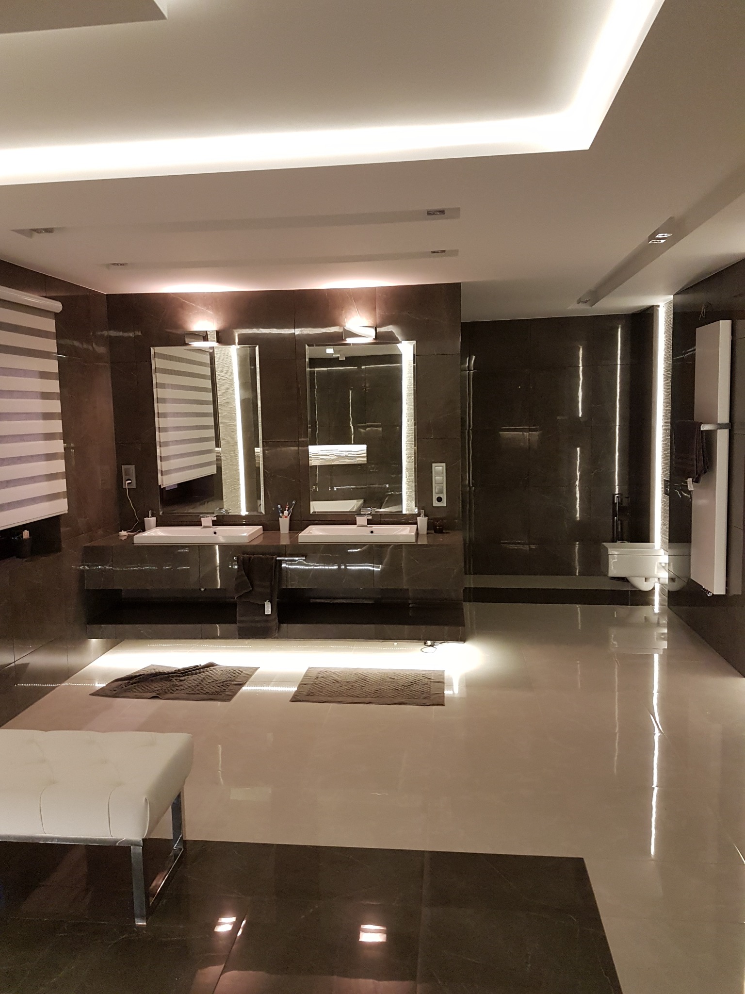 Luksusowa łazienka z marmurowymi płytkami, podwójną umywalką z podświetlanymi lustrami i nowoczesnym oświetleniem sufitowym. Widoczny biały puf i grzejnik.