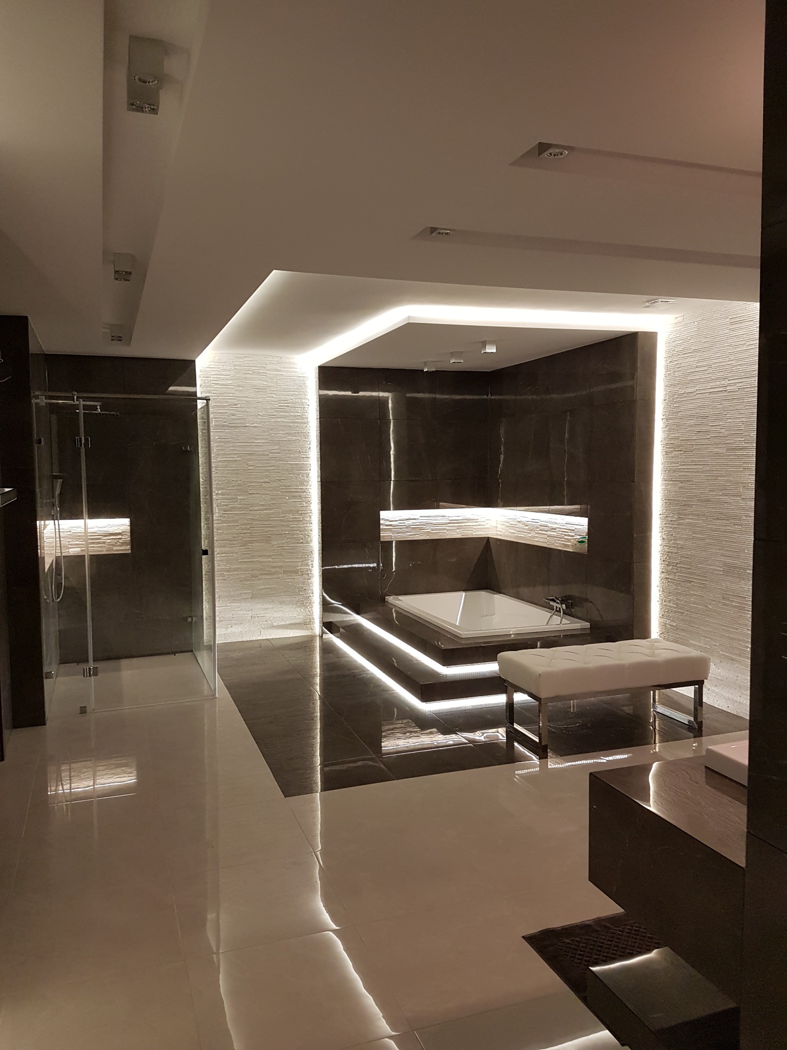 Nowoczesna łazienka z kabiną prysznicową, wanną i podświetleniem LED. Ciemne płytki kontrastują z białą armaturą i teksturowaną ścianą. Minimalistyczny design.