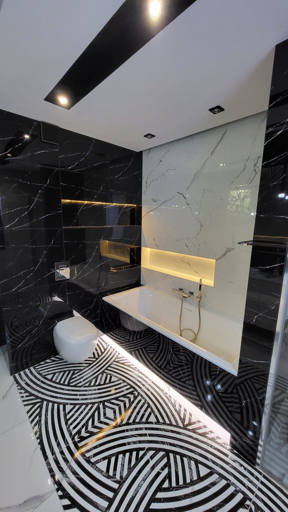 Nowoczesna łazienka z czarno-białymi płytkami imitującymi marmur, podświetlanymi niszami i wzorzystą podłogą. Uwagę przykuwa geometryczny wzór płytek podłogowych.
