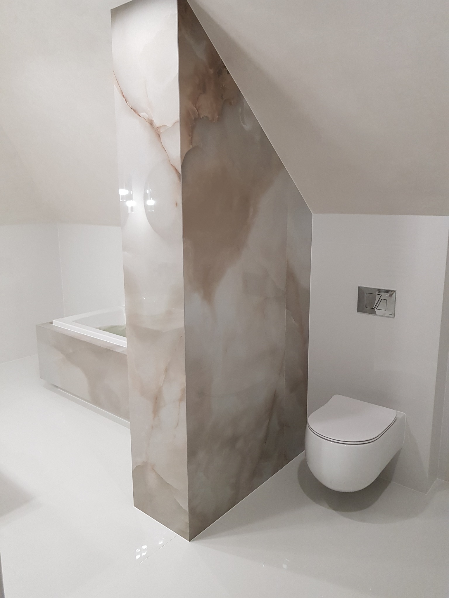Nowoczesna łazienka na poddaszu z marmurową ścianką działową, podwieszaną toaletą i wanną w zabudowie. Minimalistyczny design, jasne płytki, skośny sufit.