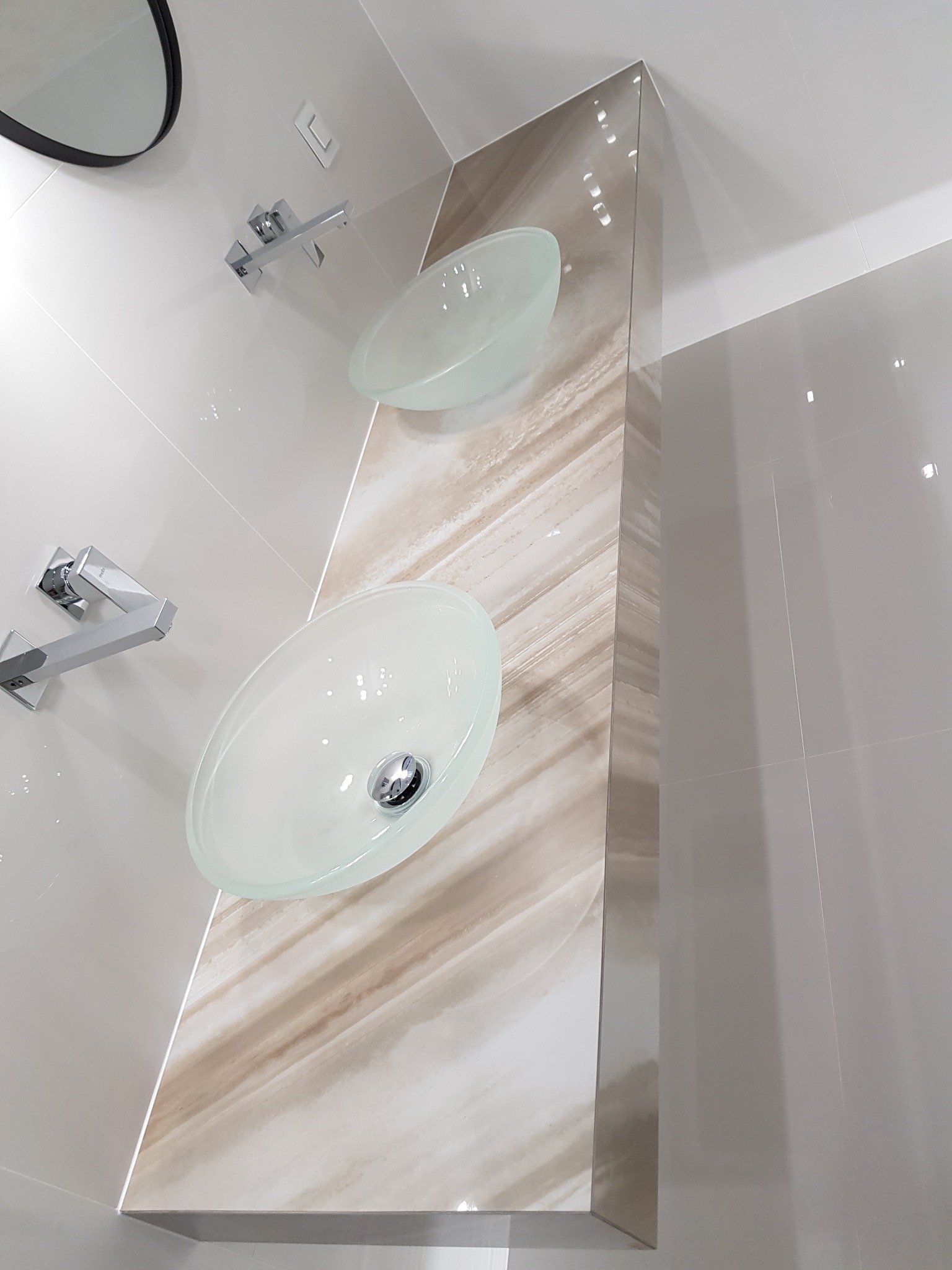 Dwie szklane umywalki nablatowe z chromowanymi bateriami na ścianie wyłożonej beżowymi płytkami i dekoracyjnym panelem imitującym marmur. Nowoczesna łazienka.