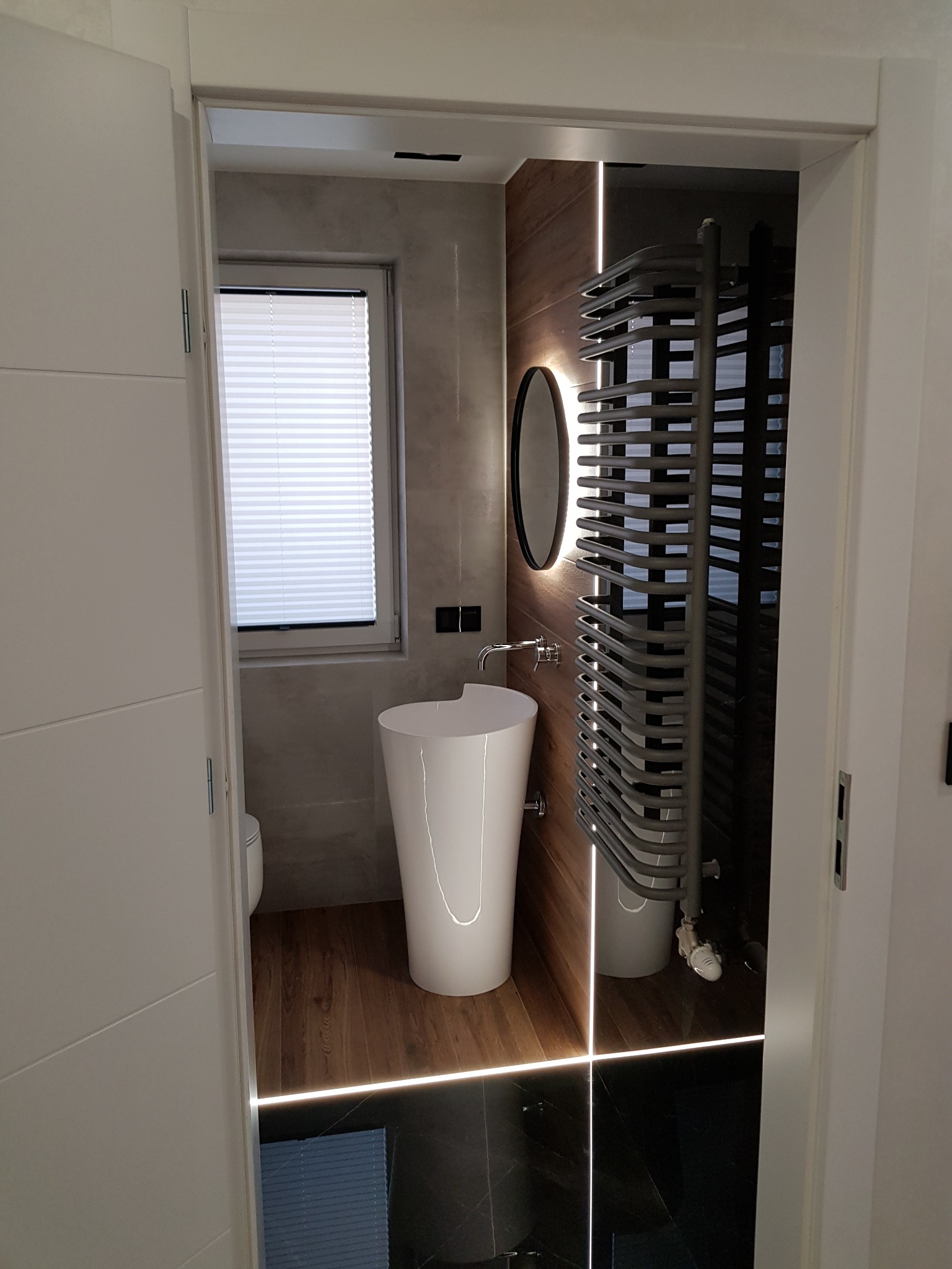 Nowoczesna łazienka z wolnostojącą umywalką, podświetlanym lustrem i grzejnikiem drabinkowym. Kontrastujące płytki podłogowe i drewniane akcenty.