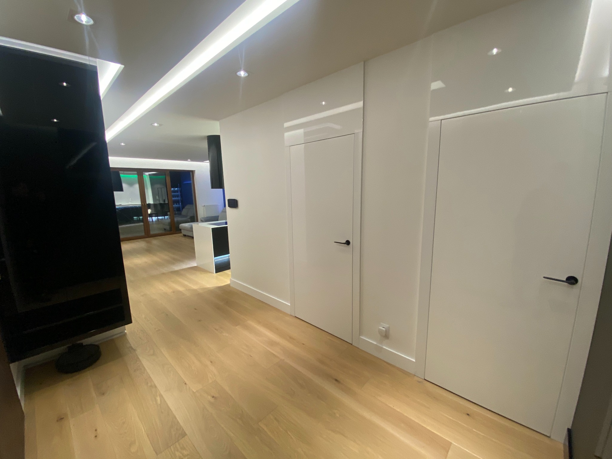 Nowoczesne wykończenie wnętrza: jasna podłoga, białe ściany z ukrytymi drzwiami i minimalistyczne oświetlenie. Widoczny fragment salonu w głębi.