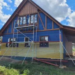 Dom w trakcie ocieplania, widoczne rusztowanie, niebieska membrana wiatroizolacyjna i żółta wełna mineralna, drewniana konstrukcja dachu, błękitne niebo z chmurami w tle.