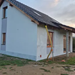 Surowy tynk elewacyjny na nowym domu jednorodzinnym, widoczne rury wentylacyjne, okna zabezpieczone folią, przygotowanie do dalszych prac wykończeniowych.